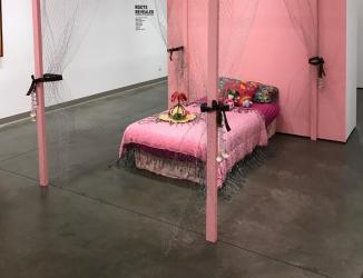 Image of installation artwork by Gabrielle Moore depicting a pink tropical themed bed setting, surrounded by a wire curtains and barbed wire embelishments.