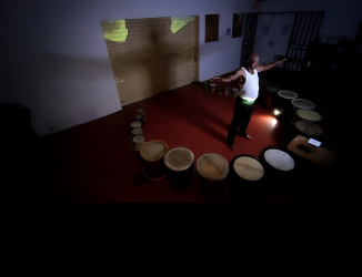 Image capture showing a man standing in the middle of a round of drums of various sizes, from the experimental film 'I dare, I promise'