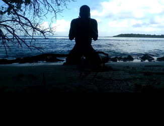 Image capture showing a man sitting on the ground under a tree and with his back to the sea on the shore of a deserted beach, from the experimental film 'I dare, promise'