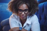 Photograph of a mixed race Black woman with an afro with blonde highlights and golden hair wearing black glasses smiling at the camera